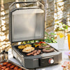 A portable Royal Gourmet electric grill with a hinged lid, shown open and cooking chicken, steak, and vegetables on a patio table. The grill has a dark gray body with a silver interior and a red control dial.