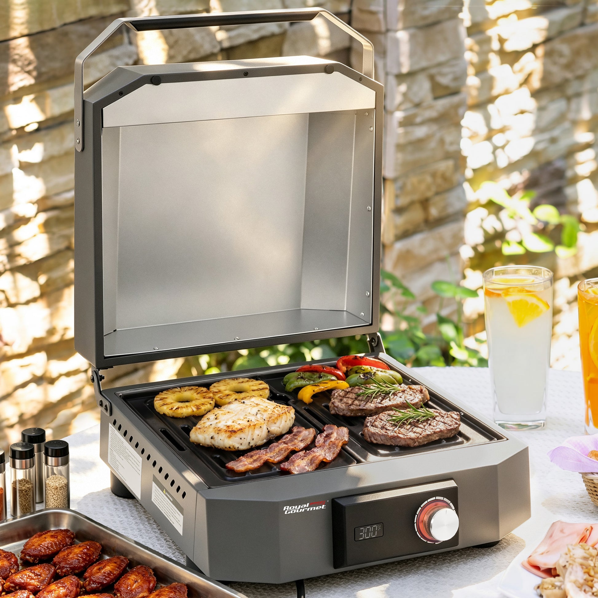 A portable Royal Gourmet electric grill with a hinged lid, shown open and cooking chicken, steak, and vegetables on a patio table. The grill has a dark gray body with a silver interior and a red control dial.