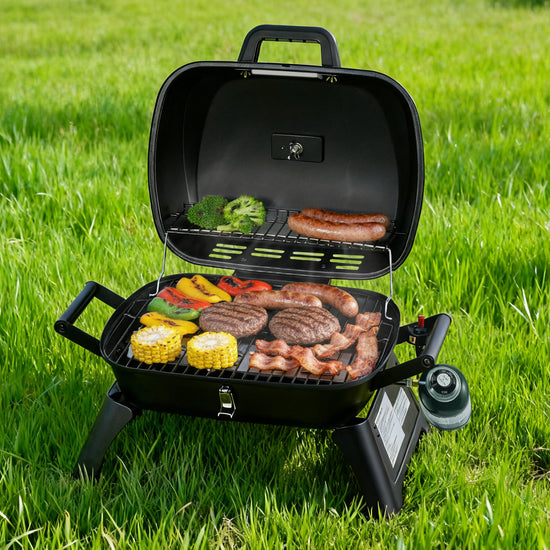 A black Royal Gourmet portable gas grill with its lid open, shown outdoors on green grass while cooking sausages, burgers, corn, and mixed vegetables.