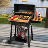 A black Royal Gourmet charcoal grill with a wooden lid handle and side table is set on a wooden deck overlooking a lake. The grill is open, showing cooked meat, vegetables, and skewers, with bottles of sauce on the side table and bags of charcoal underneath.