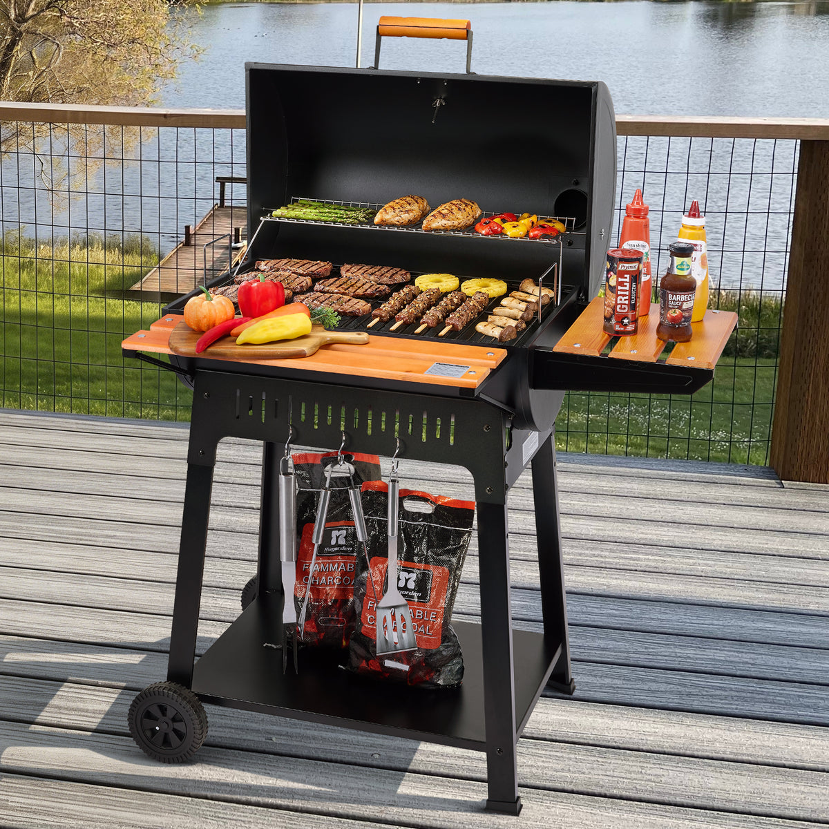 A black Royal Gourmet charcoal grill with a wooden lid handle and side table is set on a wooden deck overlooking a lake. The grill is open, showing cooked meat, vegetables, and skewers, with bottles of sauce on the side table and bags of charcoal underneath.