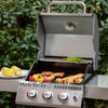 A stainless steel Royal Gourmet gas grill with the lid open, cooking sausages, chicken, peppers, and asparagus outdoors. A plate of corn is on the left, with leafy greenery in the background.
