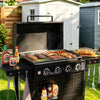 A black outdoor Royal Gourmet gas grill with the lid open, cooking burgers, sausages, steak, and tomatoes on a flat griddle tray. The grill is set on a grassy lawn in a backyard with a white shed and wooden fence in the background.
