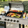 A stainless steel Royal Gourmet gas grill outdoors on a lawn, loaded with grilled meat, vegetables, and a pot of soup on its burners. Condiment bottles sit on the side, and the grill lid is open to show the cooking food.