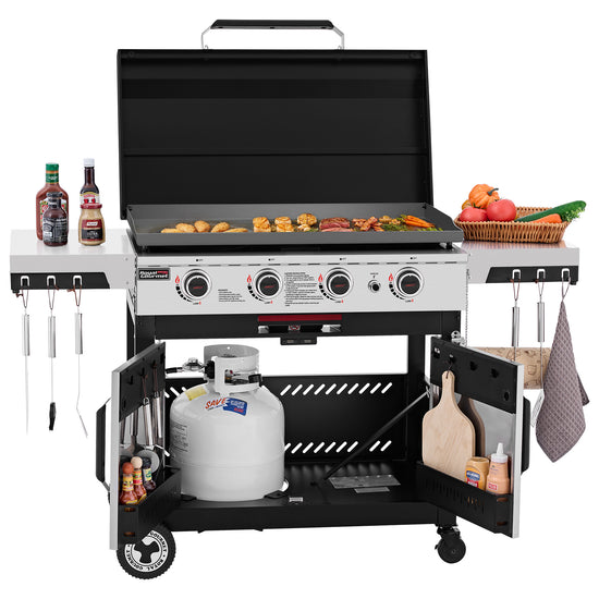 A large black and silver Royal Gourmet gas grill with the lid open, cooking food on the grates. The grill has side shelves holding condiments and vegetables, hanging utensils, a cutting board, and a propane tank in the lower compartment, all shown on a white background.