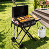 A black outdoor Royal Gourmet gas grill with wheels is set on a green lawn, cooking burgers, steak, corn, and vegetables. A white propane tank is connected to the grill, and a wooden deck with patio chairs is visible in the background.