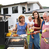 A family of four enjoys outdoor grilling in a backyard setting. An older woman in a blue and white striped top serves food from a black Royal Gourmet charcoal grill, while a young woman in a red top holds a baby.