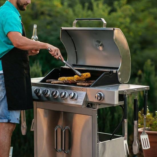 A man grilling outdoors next to a Royal Gourmet stainless steel gas barbecue grill, turning sausages and corn on the grate. The man is wearing a turquoise shirt, black apron, and denim shorts, holding a beer bottle and tongs.