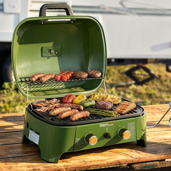 An outdoor green portable Royal Gourmet gas grill with its lid open, cooking sausages, burgers, peppers, and asparagus on a wooden table. The grill features two control knobs and the 'Grillmaster' brand logo on the front.