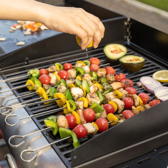 Grilled vegetable and chicken skewers on a black Royal Gourmet  outdoor grill, with a hand squeezing lemon juice over the food. Sides include sliced red onion, avocado, and lemon wedges.