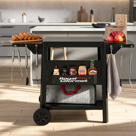 A black Royal Gourmet rolling cart with wheels, shown in a modern kitchen setting. The cart has two shelves, with the top shelf holding a wooden cutting board with fried foods and a paper bag of apples, and the bottom shelf holding condiments and a gray storage bag.