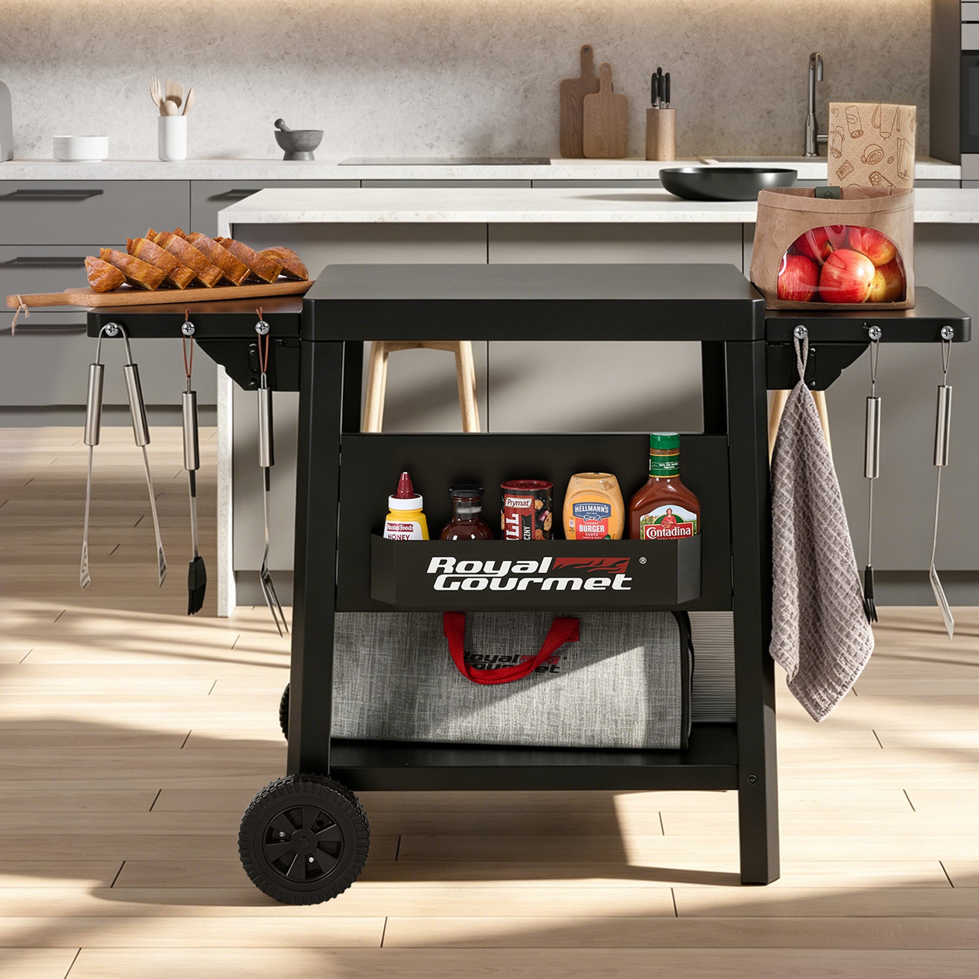 A black Royal Gourmet rolling cart with wheels, shown in a modern kitchen setting. The cart has two shelves, with the top shelf holding a wooden cutting board with fried foods and a paper bag of apples, and the bottom shelf holding condiments and a gray storage bag.