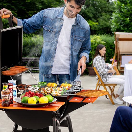 A man grilling outdoors at a patio party, standing behind a black charcoal grill with an orange wood-burning side table attached. The table holds lemons, limes, tomatoes, peppers, and a platter of mixed vegetables.