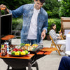 A man grilling outdoors at a patio party, standing behind a black charcoal grill with an orange wood-burning side table attached. The table holds lemons, limes, tomatoes, peppers, and a platter of mixed vegetables.
