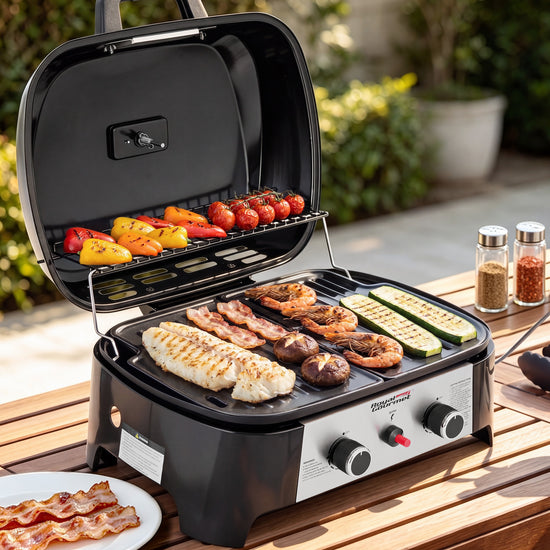 A black and silver Royal Gourmet portable electric grill sits open on a wooden outdoor table, cooking fish, bacon, shrimp, zucchini, and mushrooms. Colorful peppers and tomatoes are grilling on the upper rack.