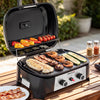 A black and silver Royal Gourmet portable electric grill sits open on a wooden outdoor table, cooking fish, bacon, shrimp, zucchini, and mushrooms. Colorful peppers and tomatoes are grilling on the upper rack.