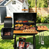 A black Royal Gourmet  outdoor charcoal grill with a side table and hanging utensils, shown fully loaded with steak, corn, asparagus, peppers, and sausages on a grassy backyard lawn. A bag of charcoal and cooler bag hang below the grill.