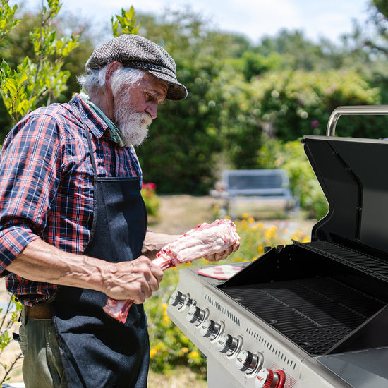 An older man with a white beard, wearing a plaid shirt, dark apron, and tweed cap, is grilling a bone-in cut of meat next to a Royal Gourmet stainless steel gas grill in an outdoor garden setting.