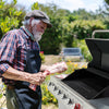 An older man with a white beard, wearing a plaid shirt, dark apron, and tweed cap, is grilling a bone-in cut of meat next to a Royal Gourmet stainless steel gas grill in an outdoor garden setting.