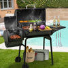 A black Royal Gourmet  outdoor charcoal grill with side tables and hanging utensils, shown on a lawn by a swimming pool with food cooking inside and condiments on the side deck. The grill has a visible 'Bourgeo' brand bag hanging from the lower shelf.