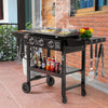 A black Royal Gourmet outdoor gas grill cart with wheels, shown on a patio with food cooking on the griddle, bottles on the shelf, and utensils hanging on the sides.