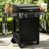 A black outdoor Royal Gourmet gas grill on a wheeled cart with side shelves, shown in a backyard setting. The grill has blue control knobs, a digital temperature display, and accessories including utensils, bottles, and a basket of bread on its side tables.
