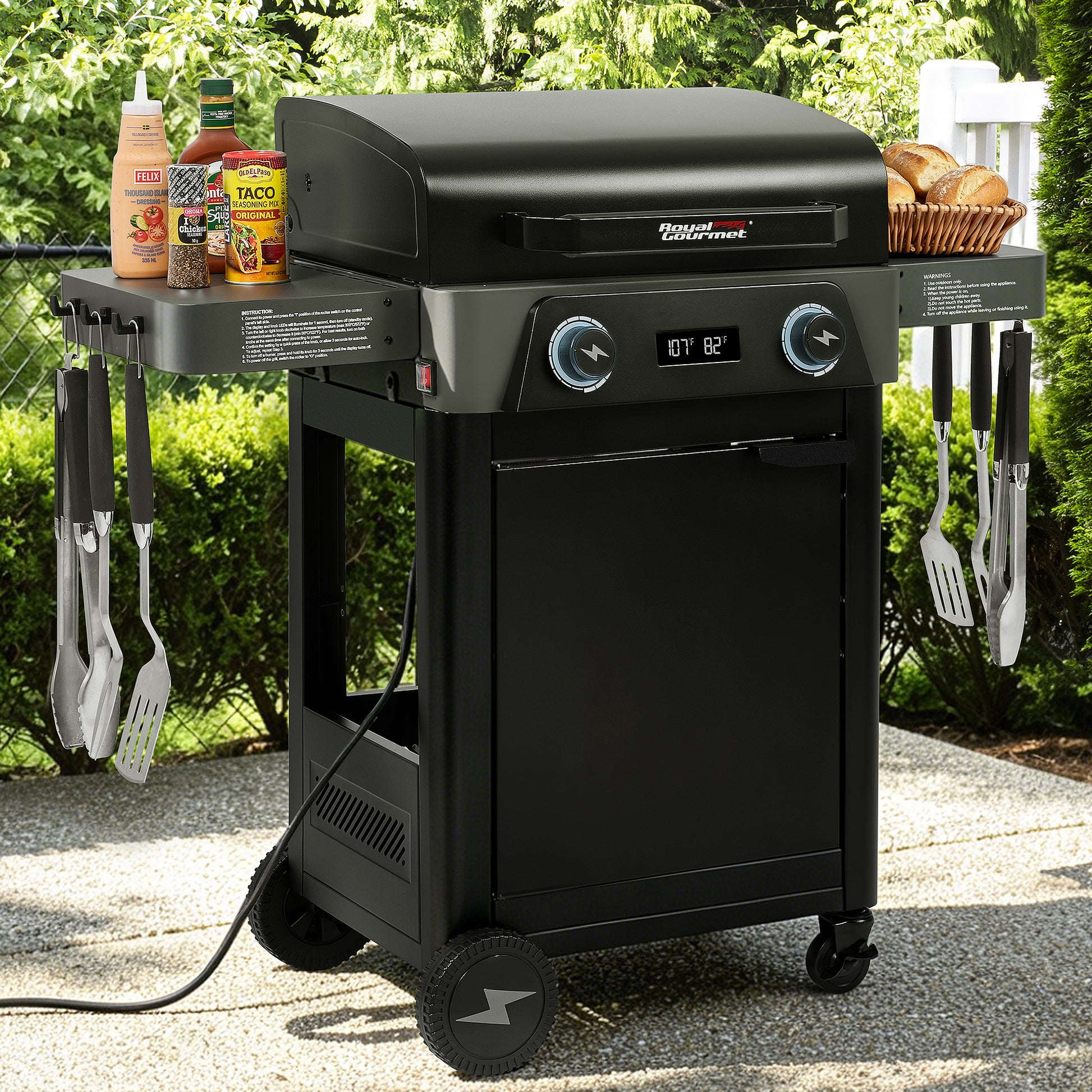 A black outdoor Royal Gourmet gas grill on a wheeled cart with side shelves, shown in a backyard setting. The grill has blue control knobs, a digital temperature display, and accessories including utensils, bottles, and a basket of bread on its side tables.