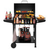 A black Royal Gourmet charcoal grill with a lid, shown open and loaded with food on the grates and side shelves, set against a plain white background. Condiments, charcoal bags, a towel, and a spatula are arranged around the grill.