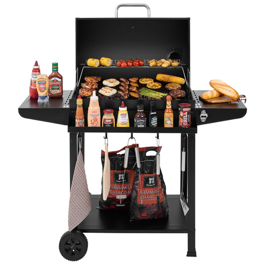 A black Royal Gourmet charcoal grill with a lid, shown open and loaded with food on the grates and side shelves, set against a plain white background. Condiments, charcoal bags, a towel, and a spatula are arranged around the grill.
