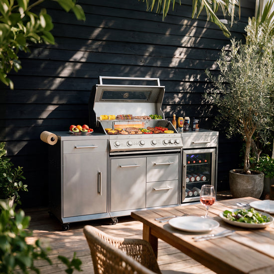 A stainless steel Royal Gourmet outdoor kitchen set with a built-in grill, wine cooler, and side cabinets, staged on a wooden deck against a dark wood-paneled wall with potted plants and a dining table in the foreground.