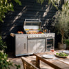 A stainless steel Royal Gourmet outdoor kitchen set with a built-in grill, wine cooler, and side cabinets, staged on a wooden deck against a dark wood-paneled wall with potted plants and a dining table in the foreground.