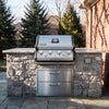A stainless steel outdoor Royal Gourmet  gas grill with four burners is mounted on a stone and concrete backyard island, with a brick house and evergreen trees in the background.