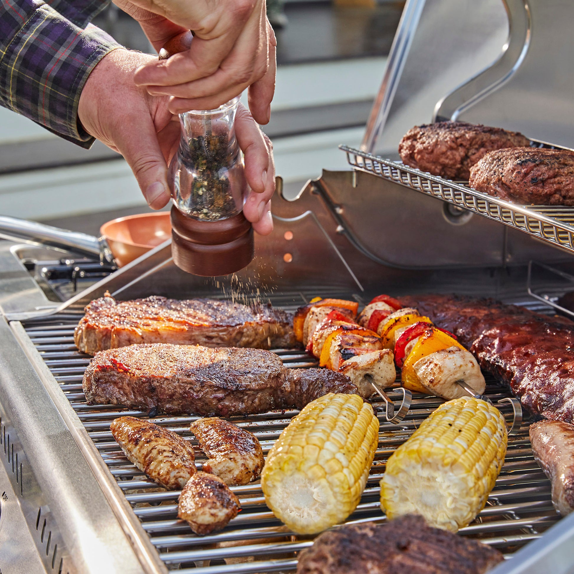 A close-up lifestyle shot of a person seasoning a stainless steel Royal Gourmet outdoor grill with a glass pepper grinder. Freshly grilled steaks, corn on the cob, chicken skewers, and burgers are cooking on the grill grates.