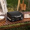Black portable Royal Gourmet grill on a wooden table outdoors with a tent and cutting board in the background.