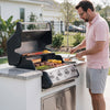 A man in a pink polo shirt and gray shorts grilling outdoors on a stainless steel and black Royal Gourmet  gas grill built into a white outdoor kitchen island with a granite countertop.