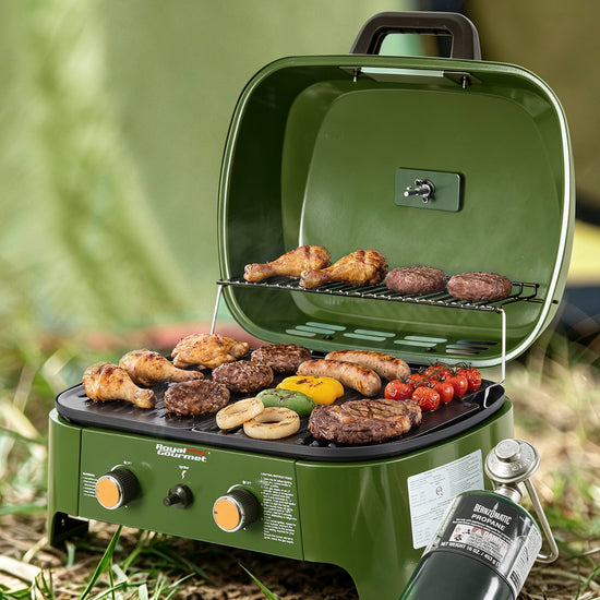 An Royal Gourmet outdoor portable camping grill in olive green, shown open with food cooking on the grates. The grill has two control knobs on the front, a carrying handle, and a propane tank attached.