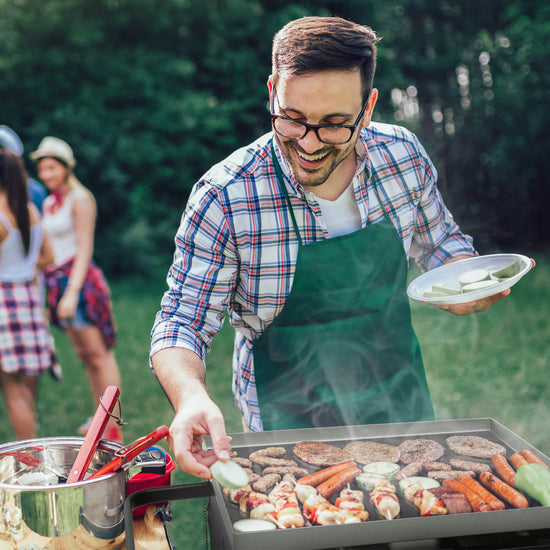 A man wearing a green apron and glasses grills burgers and sausages outdoors on a hot day, holding a plate of sliced cucumbers. Other people are blurred in the background on a grassy lawn.