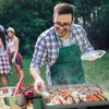 A man wearing a green apron and glasses grills burgers and sausages outdoors on a hot day, holding a plate of sliced cucumbers. Other people are blurred in the background on a grassy lawn.
