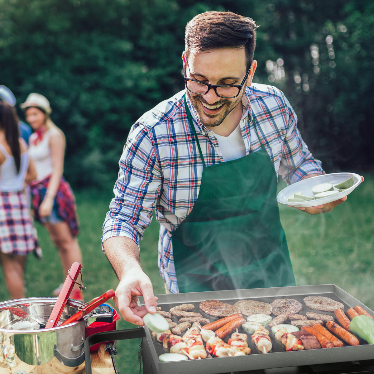 A man wearing a green apron and glasses grills burgers and sausages outdoors on a hot day, holding a plate of sliced cucumbers. Other people are blurred in the background on a grassy lawn.