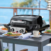 Black Royal Gourmet portable grill on a table with food and a cup, set against a blurred outdoor background.