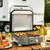 A compact electric Royal Gourmet  grill with a silver interior and gray exterior sits on a wooden picnic table outdoors, cooking skewers, bacon, potatoes, and asparagus. A bottle of beer, food plates, and a white RV are visible in the background.