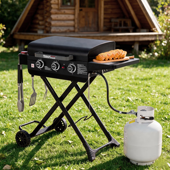 A black Royal Gourmet portable folding grill with side accessories is set on a green lawn in front of a wooden log cabin. A white propane tank is connected to the grill, and a wooden board with sausages sits on the side tray.