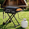 A black Royal Gourmet portable folding grill with side accessories is set on a green lawn in front of a wooden log cabin. A white propane tank is connected to the grill, and a wooden board with sausages sits on the side tray.