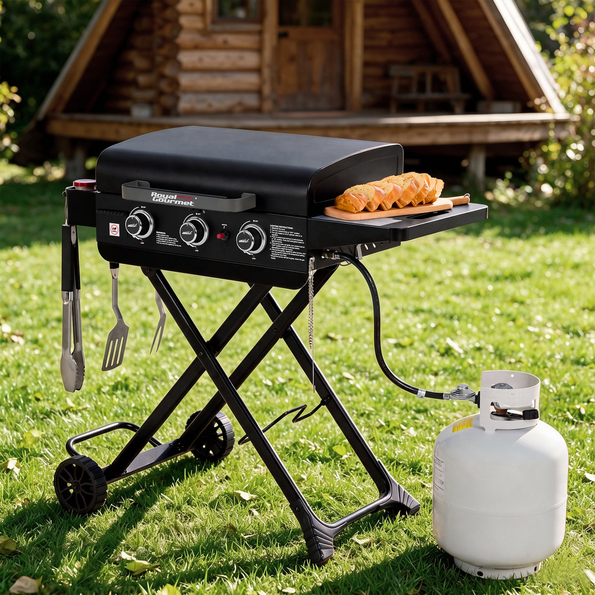 A black Royal Gourmet portable folding grill with side accessories is set on a green lawn in front of a wooden log cabin. A white propane tank is connected to the grill, and a wooden board with sausages sits on the side tray.
