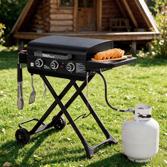 A black Royal Gourmet portable folding grill with side accessories is set on a green lawn in front of a wooden log cabin. A white propane tank is connected to the grill, and a wooden board with sausages sits on the side tray.