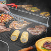 A close-up of a hand using metal tongs to turn grilled food on a black outdoor grill. Corn on the cob, steaks, shrimp, kebabs, and peppers are cooking on the grill grates, with smoke rising in the background.