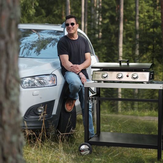 Man leaning against a car with a Royal Gourmetportable grill in a forest setting