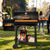 A black Royal Gourmet outdoor pellet grill with orange accents stands on a grassy lawn, with people blurred in the background and cooking accessories hanging from its side table.