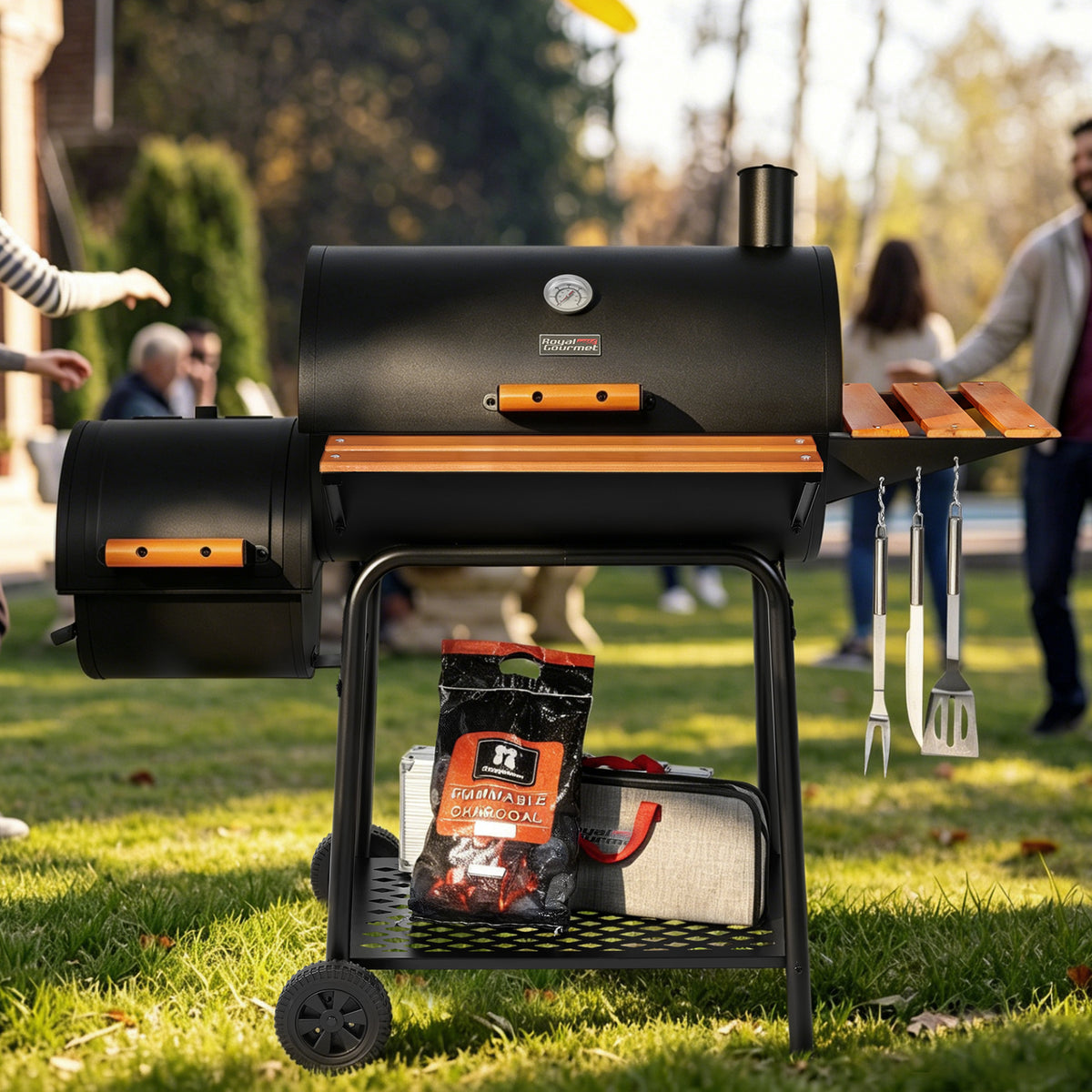 A black Royal Gourmet outdoor pellet grill with orange accents stands on a grassy lawn, with people blurred in the background and cooking accessories hanging from its side table.