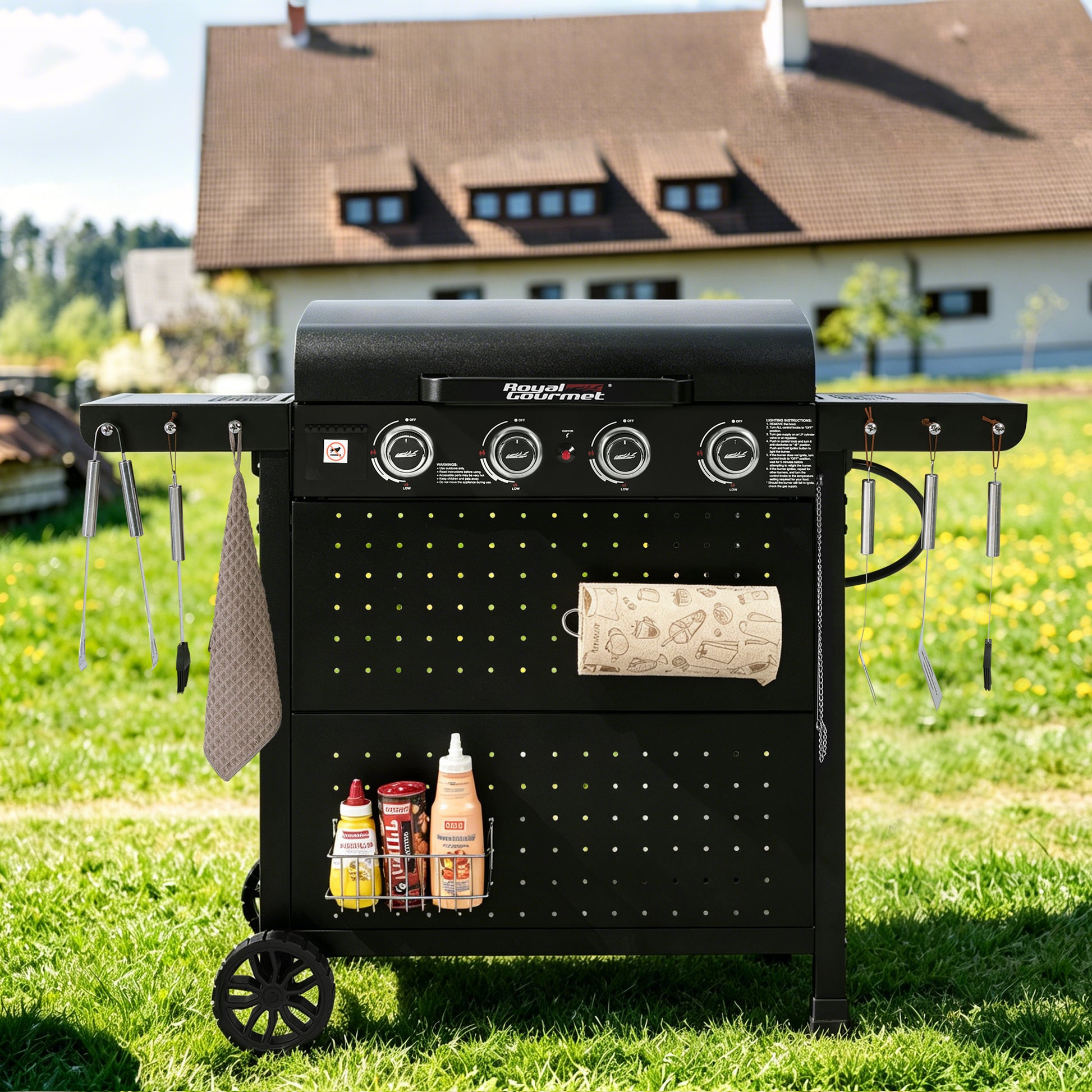 A black outdoor Royal Gourmet grill cart with side tables, hanging utensils, a paper towel holder, and a lower shelf with condiment bottles, photographed on a grassy lawn in front of a house.